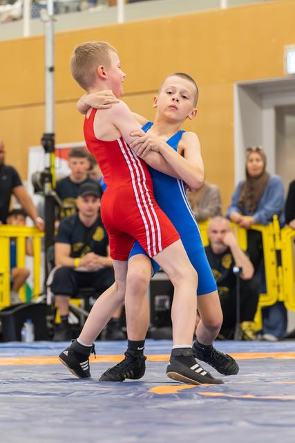 Two boys in red and blue singlets locked in a wrestling clinch, the boy in blue showing focused determination on the mat.