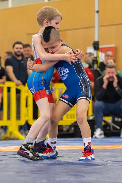 Two young wrestlers locked in a tight clinch on the mat, the boy in blue USA singlet driving forward with determination.