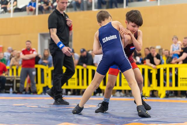 Two young boys wrestle on a blue mat; the CLReims athlete in blue grips his opponent with focused determination.