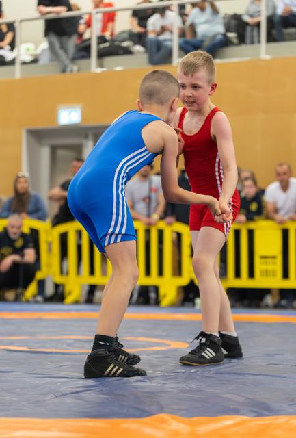Two young boys in blue and red singlets lock hands in an intense wrestling match on a blue mat, crowd watching.