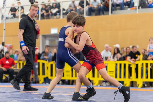 Two focused young boys wrestle on a blue mat, gripping each other intensely, referee and crowd watching behind them.