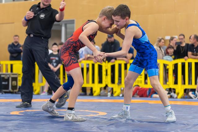 Two young wrestlers lock hands and press foreheads together in fierce concentration on a blue mat, referee watching.