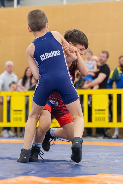 Two young wrestlers clinch in fierce grip on competition mat, blue CLReims singlet boy driving forward with intensity.