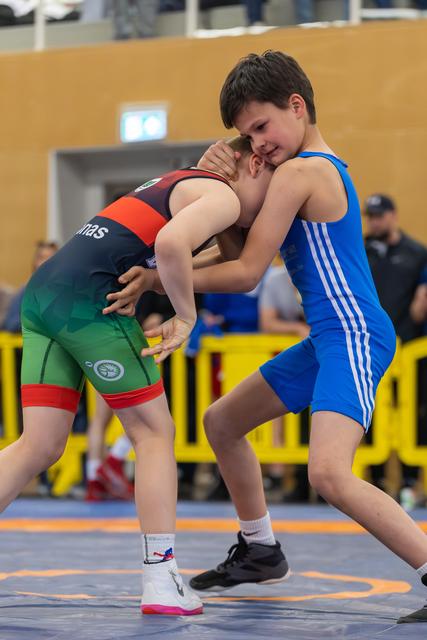 Two young wrestlers in a tight clinch; blue-suited boy smiles confidently while opponent in dark green singlet strains forward.