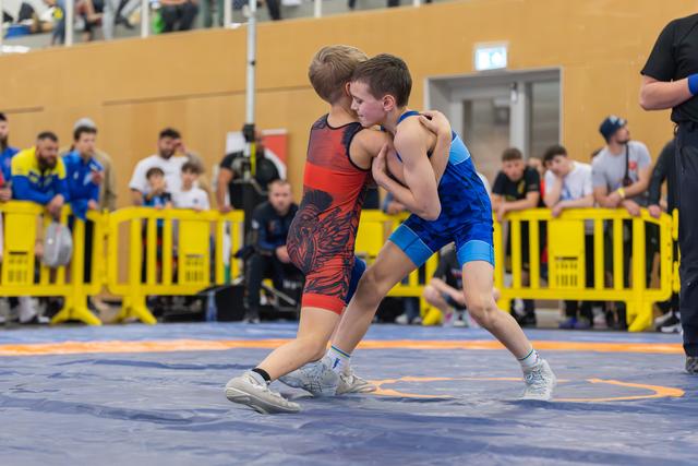 Two young boys locked in a fierce wrestling clinch on a blue mat, crowds watching behind yellow barriers.