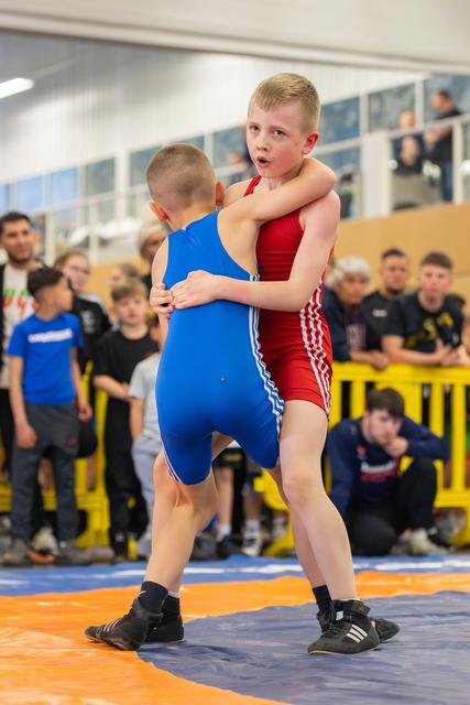 Two young boys in red and blue singlets locked in a tight clinch on the mat, crowd watching intently behind them.