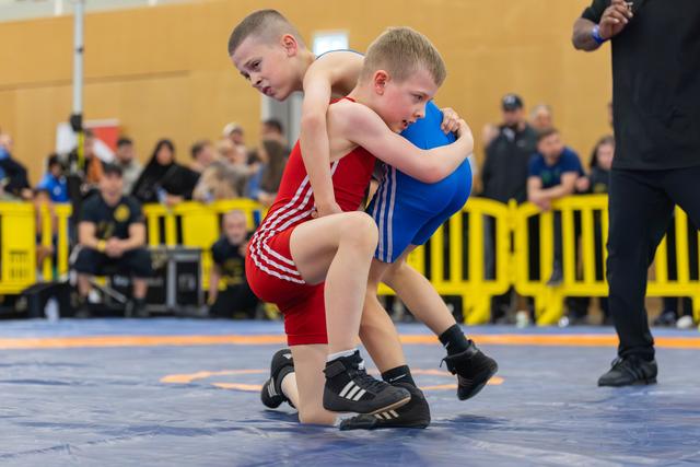 Two young boys in red and blue singlets locked in a fierce grappling clinch on a blue mat, crowd watching behind.