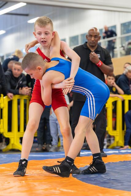 Two young boys locked in a fierce wrestling match, one in red, one in blue, with a referee watching closely behind.