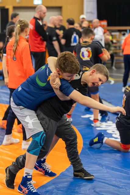 Two young boys wrestling intensely on a blue mat, one grimacing as he attempts a hold during a busy sports hall event.