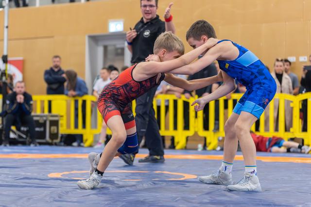 Two young boys wrestle on a blue mat, locked in a tense grip, as a referee watches closely behind them.