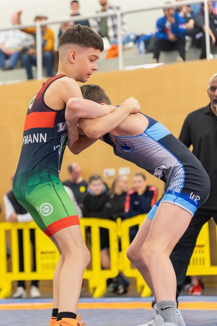 Two young wrestlers locked in a determined grip, competing indoors before a crowd of spectators.