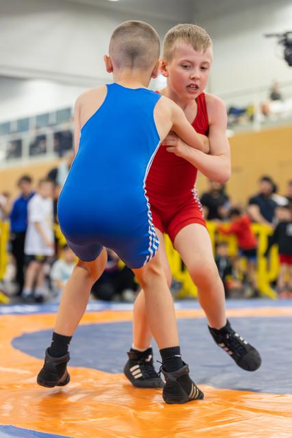 Two young boys in blue and red singlets grapple intently on an orange mat during a youth wrestling match.