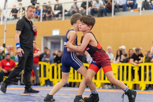 Two young boys wrestle intensely on the mat, locking grips, as a referee closely monitors nearby during a youth bout.