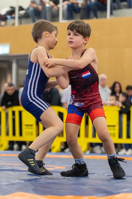 Two young wrestlers grip each other's arms in a tense standoff on the mat, both focused and determined.