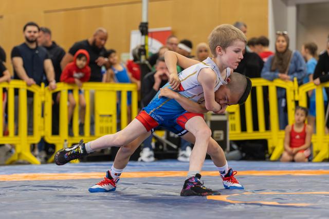 Blond boy lifts opponent in a fierce youth wrestling match on blue mats, crowd watching behind yellow barriers.