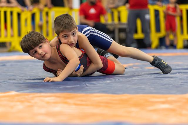 Two young boys wrestle on a blue mat, one pinning the other from behind, both showing intense focus.