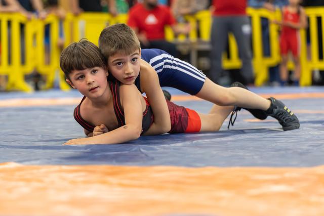 Two young boys wrestling on a blue mat, one in navy pinning the other in red, both showing intense concentration.