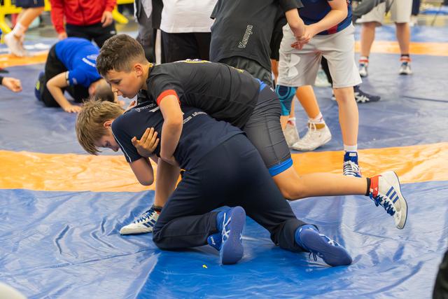 Two boys intensely wrestling on a blue mat, one applying pressure from above while others practice in the background.