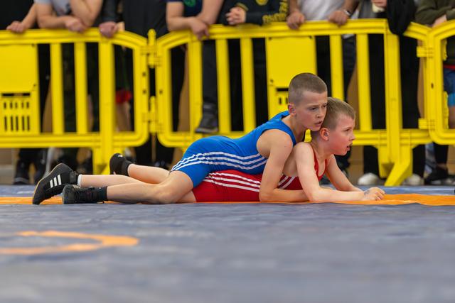 Two boys wrestle on a gray mat, one in blue controlling the other in red, both showing fierce concentration.
