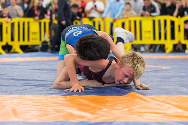 Two young boys wrestling on a blue mat, one pinning the other who strains with effort and determination.