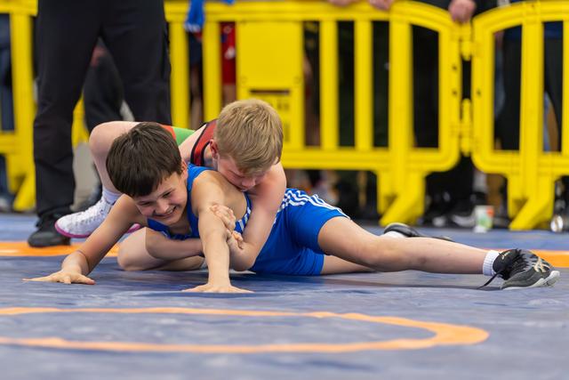 Two young boys wrestle on a blue mat, one smiling despite being pinned, showing resilience and sportsmanship.