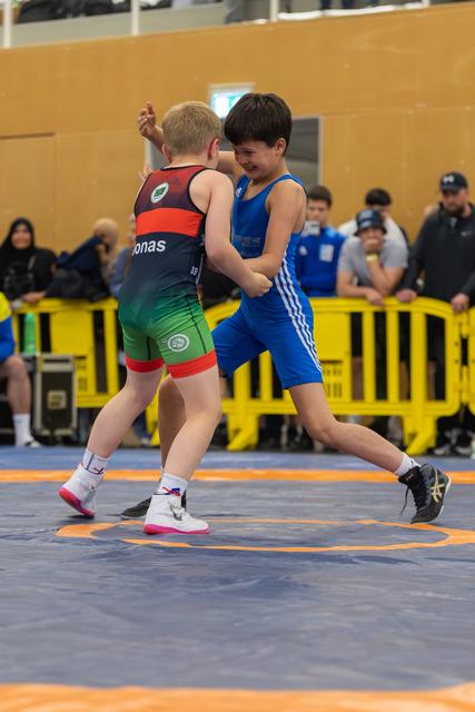 Two young boys locked in an intense wrestling bout on a blue mat, one in blue singlet lifting his leg aggressively.