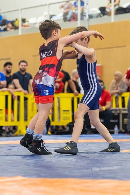 Two young boys locked in a wrestling hold on a blue mat, both focused and determined during an indoor bout.