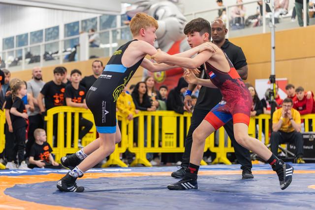 Two young boys face off on the wrestling mat, gripping each other's arms with focused intensity as a referee watches.