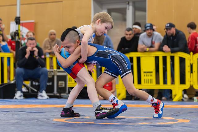 Two young boys locked in an intense wrestling hold on a blue mat, surrounded by spectators at an indoor sports event.
