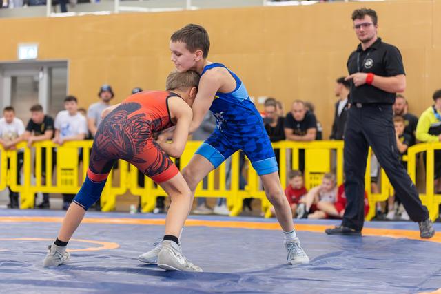 Two boys in red and blue singlets lock grips on the mat, focused and determined, as a referee watches nearby.