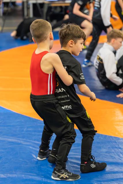 Two young boys grapple intensely on an orange and blue mat, one in red, one in black, both focused.
