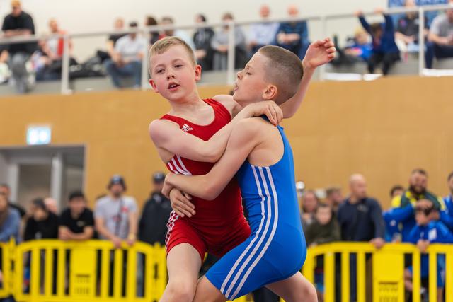 Two boys wrestle intensely in red and blue singlets, mid-grapple with determined expressions, crowd watching in background.