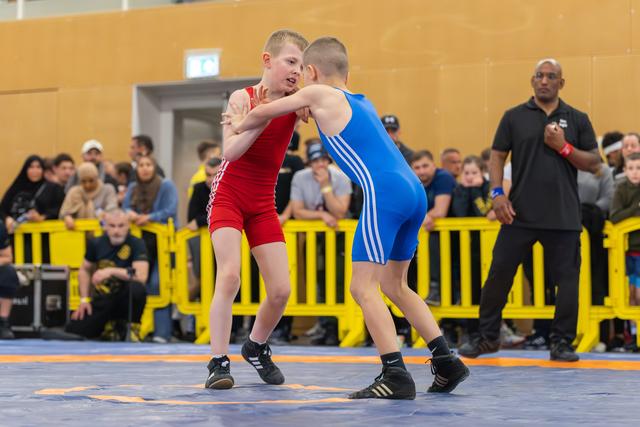 Two young wrestlers in red and blue singlets clinch intensely on the mat, watched by a referee and crowd.