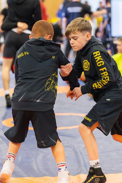 Two focused boys in RKG Freiburg hoodies wrestle intensely on a blue mat, gripping each other's arms.