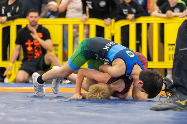 Two young boys wrestle intensely on a blue mat, one pinning the other down as spectators watch from behind yellow barriers.