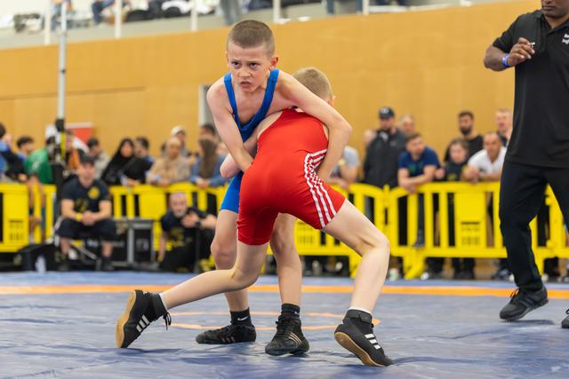Two young boys wrestle intently on a blue mat, the boy in blue grimacing with fierce concentration as he grips his opponent.