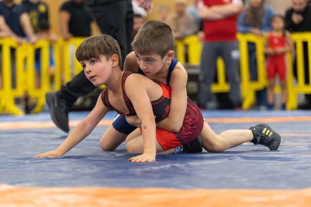 Two young boys wrestle on a blue mat, one boy controlling the other from behind, both focused and determined.