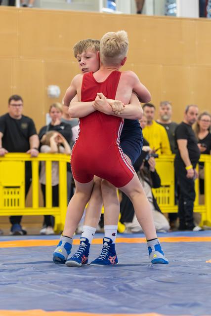 Two boys in red and blue singlets grip each other in a standing clinch, focused and determined on a blue wrestling mat.