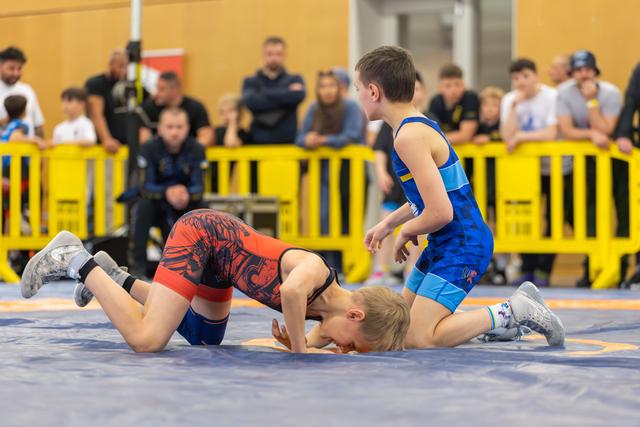 Two boys grapple on a blue mat; one in red is pinned down while the blue-suited boy stands over him, focused.