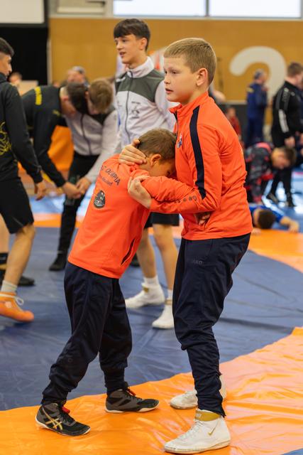 Two boys in orange jackets practice a wrestling hold on the mat, one bowing his head as the other guides firmly.