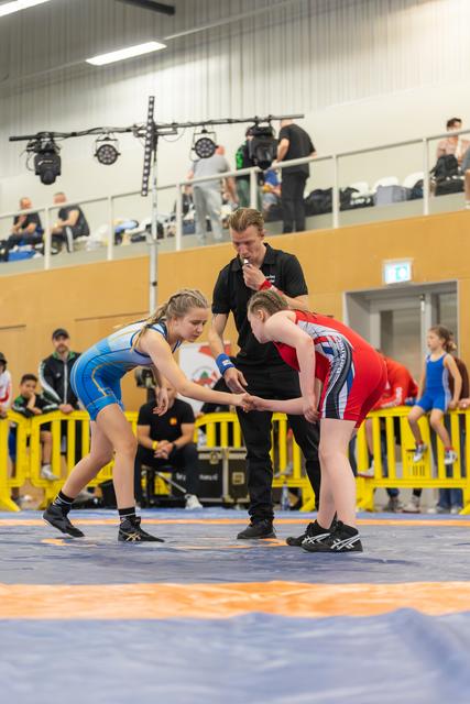 Two young female wrestlers shake hands before their bout as a referee oversees on a blue competition mat.