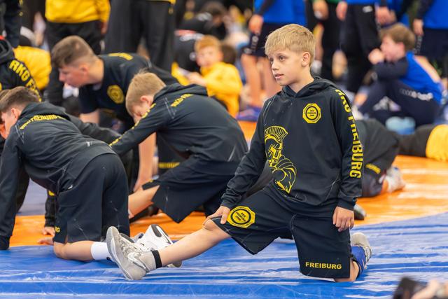 Blonde boy in Freiburg team gear stretches on the mat, looking composed while teammates warm up behind him.