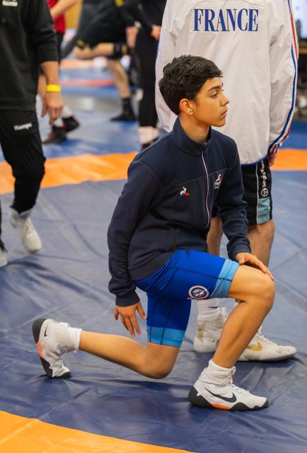 Young French wrestler performs a deep lunge stretch on the blue mat, focused and composed before competition.
