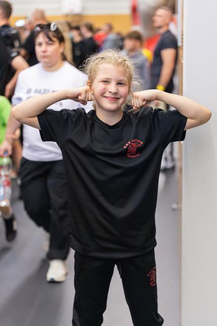 Young girl in black KAV Olympia Utrecht shirt flexes both biceps with a proud, beaming smile at a sports event.