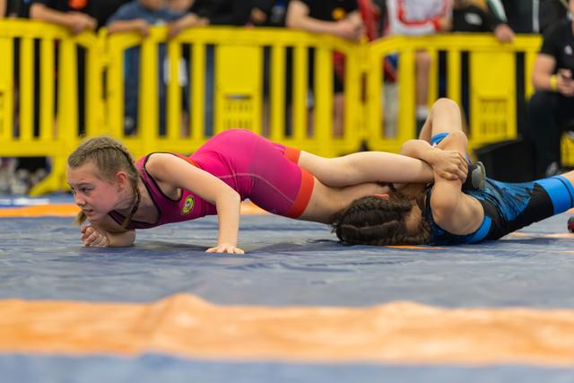 Young girl in pink singlet fights to escape a pin on the blue mat, face showing intense concentration and effort.