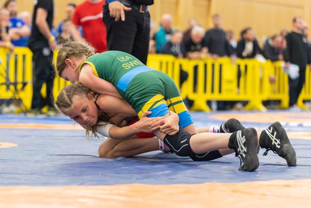 Young girl in Sweden green singlet pins opponent to blue mat, both showing intense focus during youth bout.