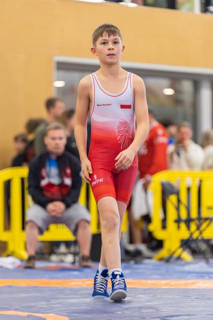 Focused young Polish wrestler walks confidently across the mat in red and white singlet, blue shoes, indoor arena.