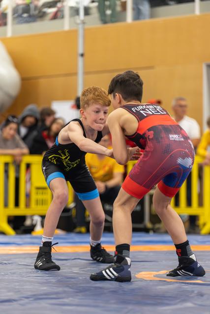 Freckled redhead boy in black singlet squares off against opponent in red NED singlet, both crouched in tense standoff.