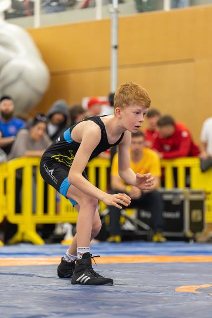 Focused young boy in wrestling singlet crouches in ready stance on blue mat, eyes alert, hands poised.