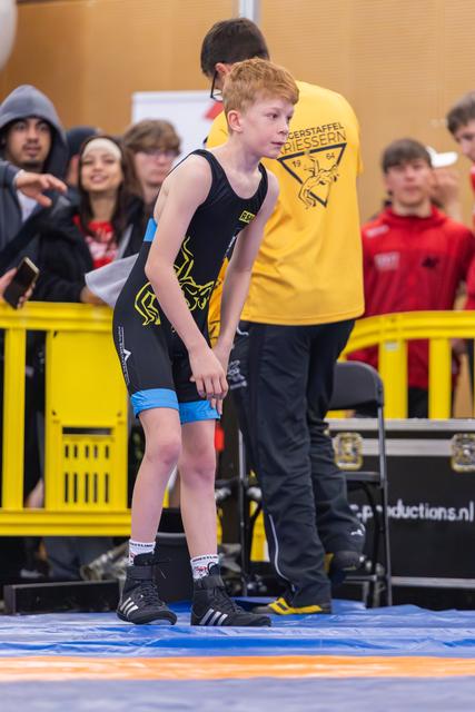 Red-haired boy in black singlet stands alert on the mat, coach in yellow behind him, crowd watching.